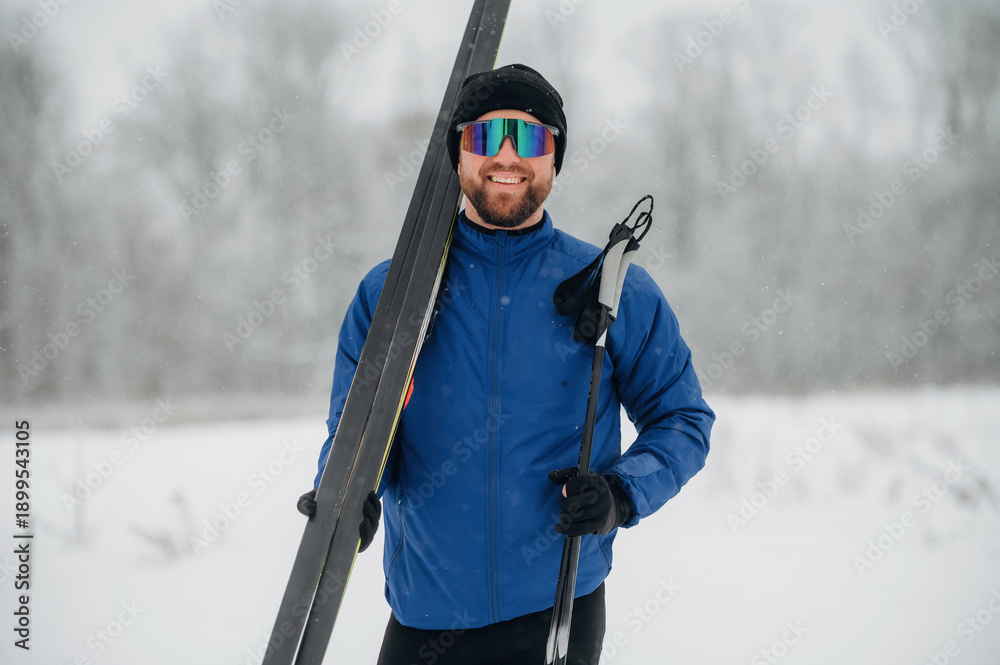 Fototapeta premium Male skier wearing blue jacket and sunglasses holds ski equipment in snowy landscape with trees and falling snowflakes in the background