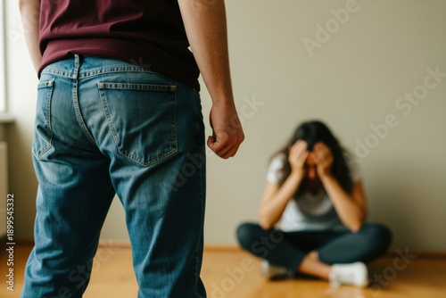 Man stands with a clenched fist in a tense indoor scene. Aggressive man faces a distressed woman sitting on the floor. Clenched fist highlights a domestic abuse situation.