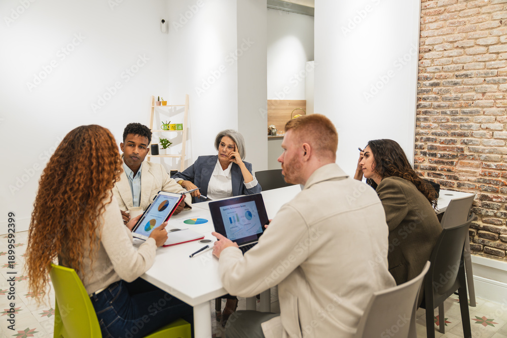 Fototapeta premium Group of diverse colleagues gathered around a table, analyzing charts and data on digital devices during a business meeting