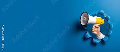 Powerful announcement and urgent message concept with hand holding a megaphone breaking through blue paper background with copy space.