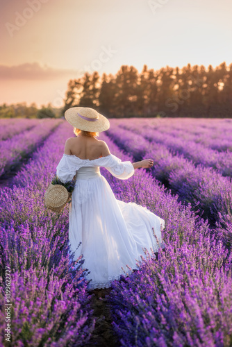 a young woman in a white dress and a big hat in a field of lavender. The view from the back, in front of the beautiful landscape of Provence and the sunset sky. In the hands of a basket with flowers