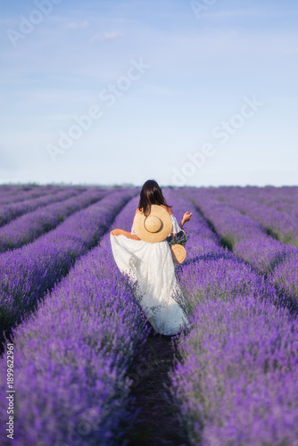 a beautiful woman in a long white dress and a large braided hat in a lavender field. View from the back. In the hands of a basket with flowers