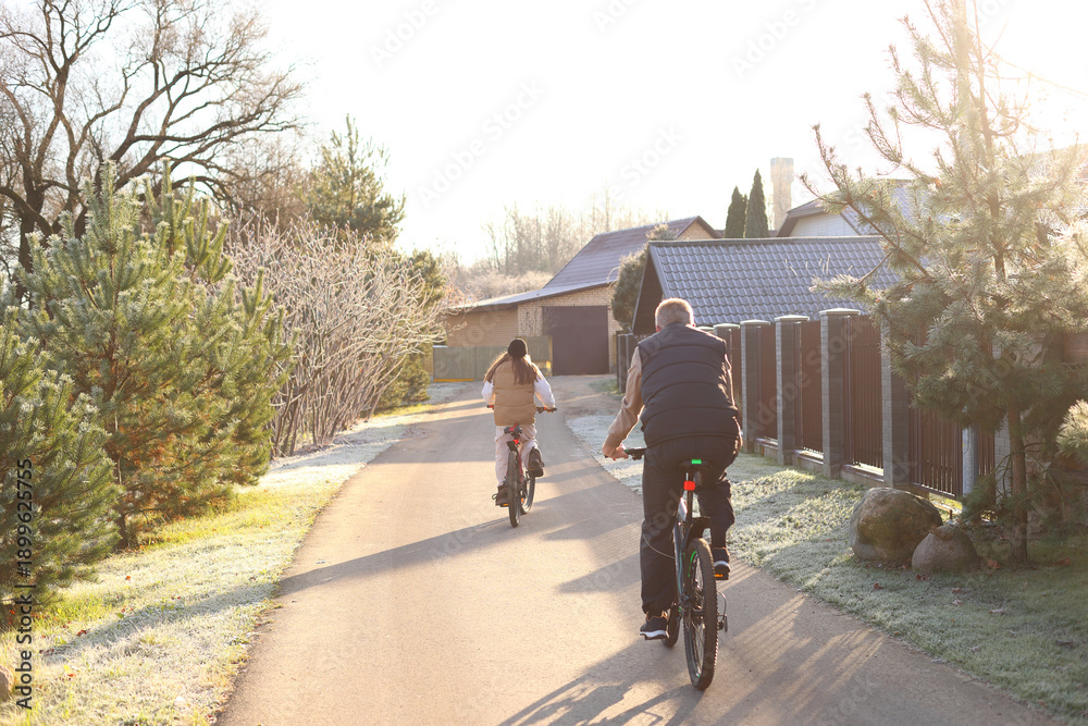Fototapeta premium A man and teen cycle away down a sunlit rural path, frosty grass, evergreens, and long shadows evoke quiet companionship, seasonal transition, and shared peaceful journey.
