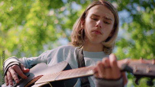 young white guitarist playing acoustic guitar in sunlit park, lowangle portrait showing focused expression, fingers on fretboard, green bokeh background, casual sweater, indie folk vibe, peaceful