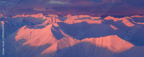 Aerial view of snow-capped peaks ablaze with the fiery hues of the setting sun, casting long, dramatic shadows across the pristine white landscape, Coldfoot, Alaska, United States.