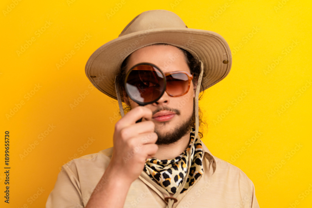 Obraz premium Man with safari hat and leopard scarf examines scene with a magnifying glass against a bright yellow background
