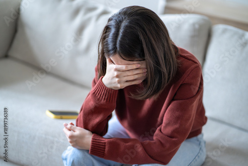 Quadro su tela Upset young woman sitting on couch covering face with hand in emotional distress, smartphone lying behind