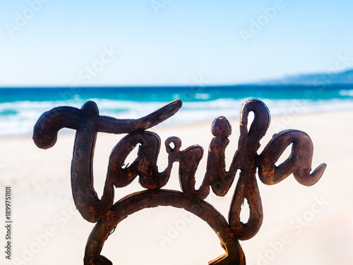 Rusty metal sign spelling “Tarifa” on a sandy beach with the Atlantic Ocean in the background