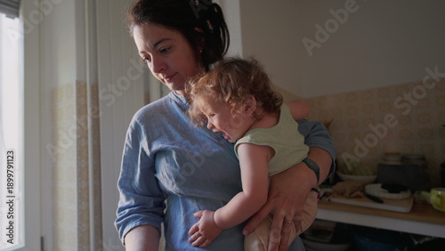 Mother holding curly-haired toddler on hip in cozy kitchen with soft window light, both gazing toward counter with gentle expression