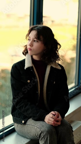 Lonely teenage girl sits alone at the window during autumn day, hugging her knees and silently thinking how to cope with problems and lack of support. Concept mental health and inner struggle of