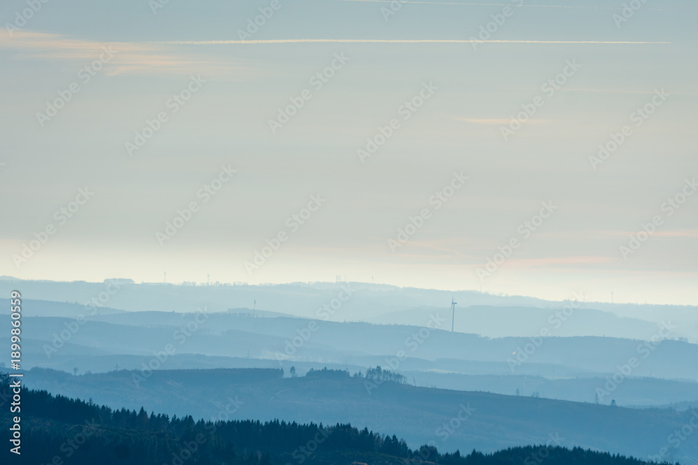 Fototapeta premium Atmospheric landscape of rolling hills gradually fading into mist under a pale sky. Soft morning light creates depth and calm mood, with wind turbines visible on the distant horizon.