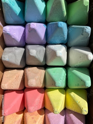 Top view of neatly arranged colorful sidewalk chalk blocks in a cardboard box. Pastel chalk pieces in soft rainbow colors create a geometric pattern with textured surfaces.