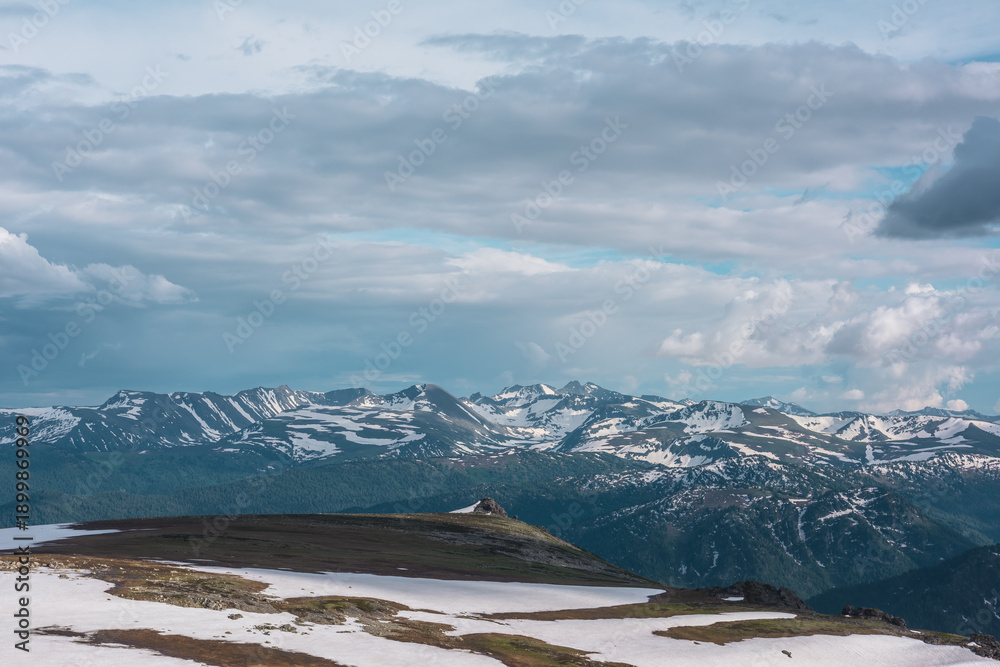 Obraz premium Snowfields and thawed ground on hill top. Sharp rocks on precipice edge above forest valley with view to big snowy mountain range far away under cloudy sky. Sunlight and shadows in high mountains.