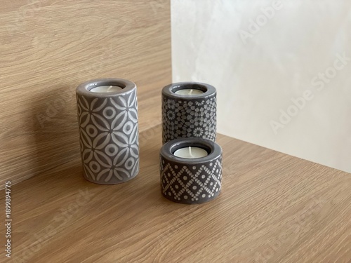 Minimalist still life of three decorative ceramic candle holders with geometric patterns placed on a wooden surface.Neutral gray tones,natural light, and simple interior background.