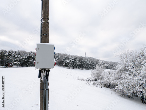 Fotografi Electrical control box mounted on a utility pole in a snowy field with forest and cloudy winter sky in the background