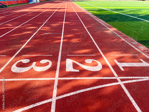 Red rubber running track lanes with start numbers 1, 2, 3 and adjacent green artificial grass field. Authentic outdoor stadium surface, ideal for racing, sports training, and competition backgrounds.