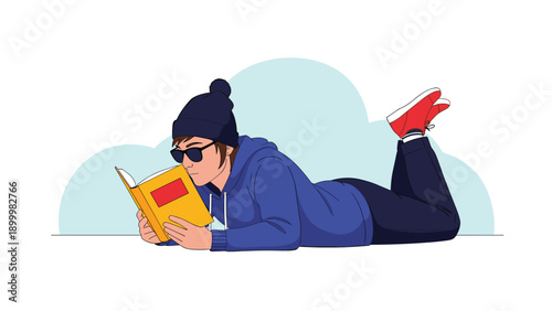 Stylish person wearing a beanie and sunglasses, comfortably lying on their stomach and engrossed in reading a book, portraying quiet leisure and learning.