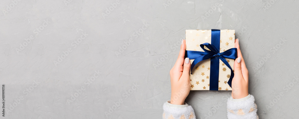 Fototapeta premium Flat lay of woman hands holding gift wrapped and decorated with bow on gray cement wooden background with copy space. Christmas and holiday concept