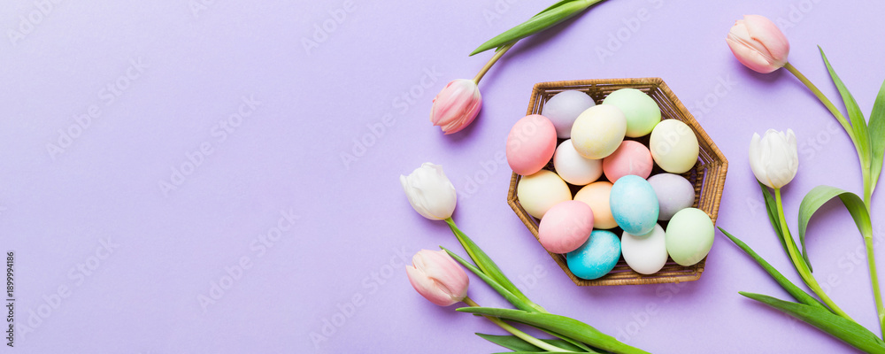 Obraz premium Happy Easter composition. Easter eggs in basket on colored table with yellow Tulips. Natural dyed colorful eggs background top view with copy space