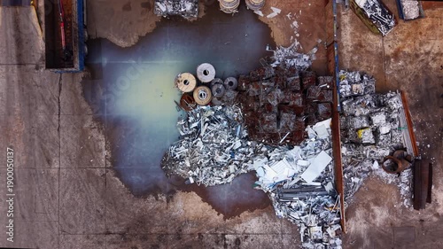 Top-down view of an urban metal scrap yard showing contaminated runoff and toxic puddles formed by industrial waste amid sorted metal stockpiles