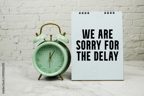 We Are Sorry For The Delay with green vintage alarm clock against a white brick wall background.