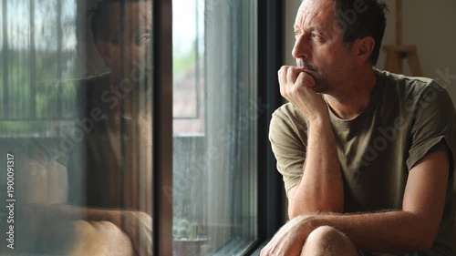 Worried middle-aged man sitting by window, resting head on hand, looking down in deep thought and sadness, symbolizing anxiety, uncertainty, and emotional isolation at home. 4K.