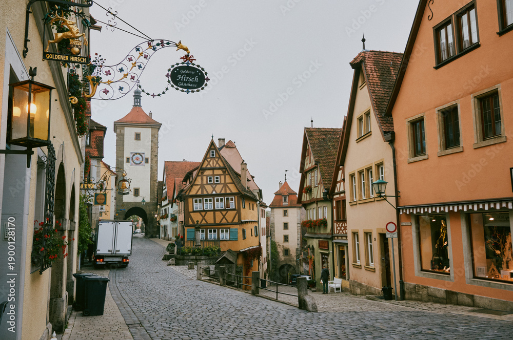 Obraz premium Cobblestone street in Rothenburg ob der Tauber lined with half-timbered houses, a clock tower, and ornate wrought-iron sign for Goldener Hirsch