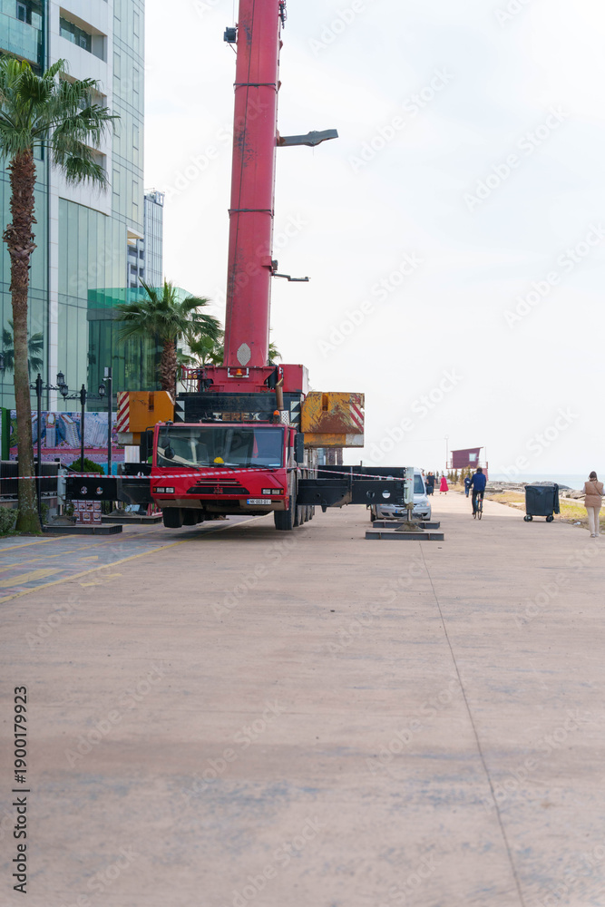 Fototapeta premium Mobile crane on waterfront promenade with safety barriers near palm trees and modern buildings