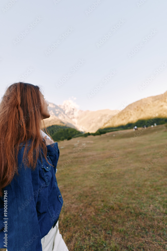 Obraz premium Woman redhead mountains meadow denim outdoors, profile portrait of young woman standing in grassy field, looking toward distant peaks at sunrise for travel, hiking and peaceful nature escape.