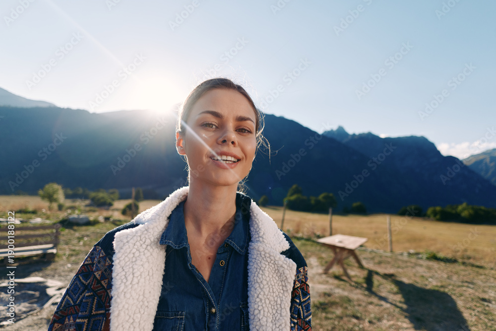 Obraz premium Woman portrait with smile against mountains outdoors at golden sunlight in countryside. Casual jacket and denim shirt, natural expression, travel lifestyle, scenic meadow background and leisure.