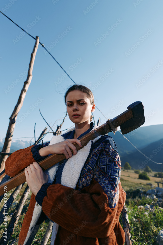 Fototapeta premium Woman with axe outdoors in rural mountains, portrait by wooden fence in a village setting, lumber tool and nature scene with rustic clothing and determined gaze.