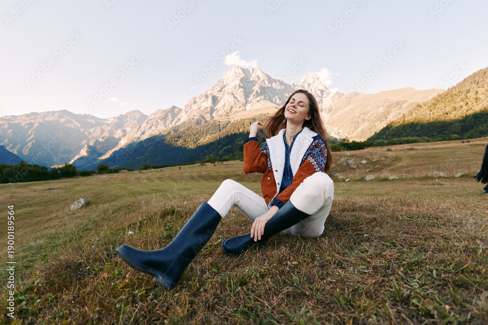 Fototapeta premium Woman sitting on mountain meadow smiling in colorful jacket and rubber boots, relaxed travel portrait in nature with distant peaks, outdoor hiking mood and warm sunlight.