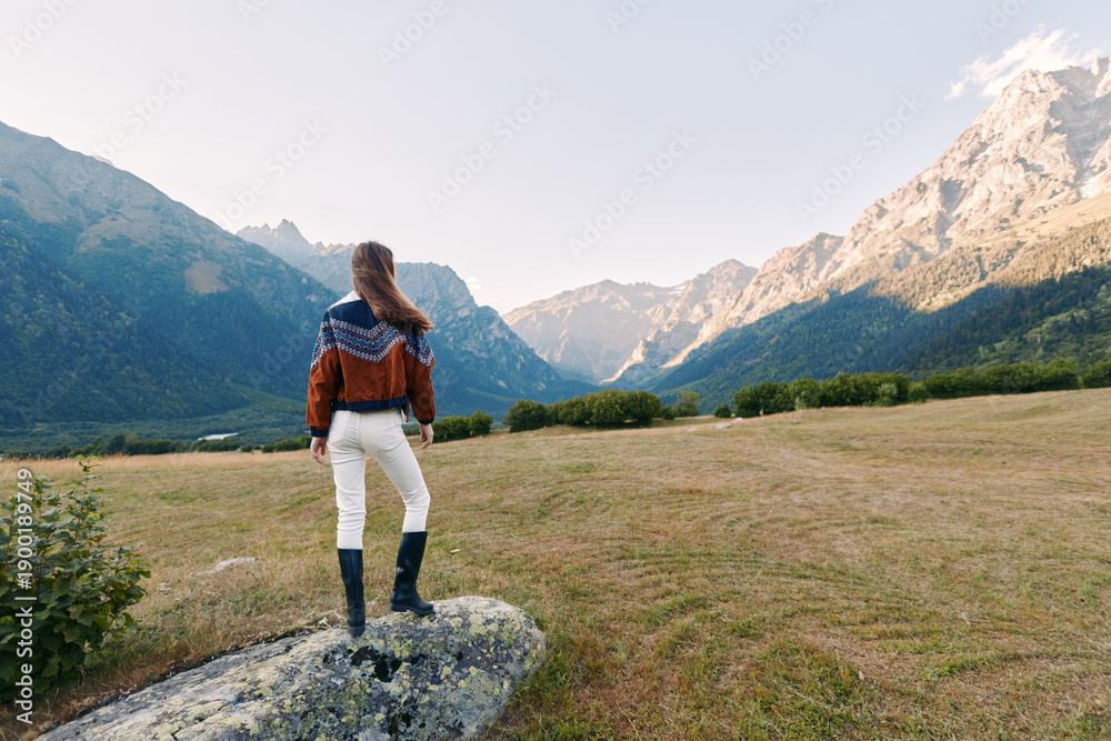 Fototapeta premium Woman stands on rock overlooking mountains landscape and nature meadow, hiking outdoors enjoying panoramic view at sunrise. Traveler in sweater and boots exploring alpine valley.