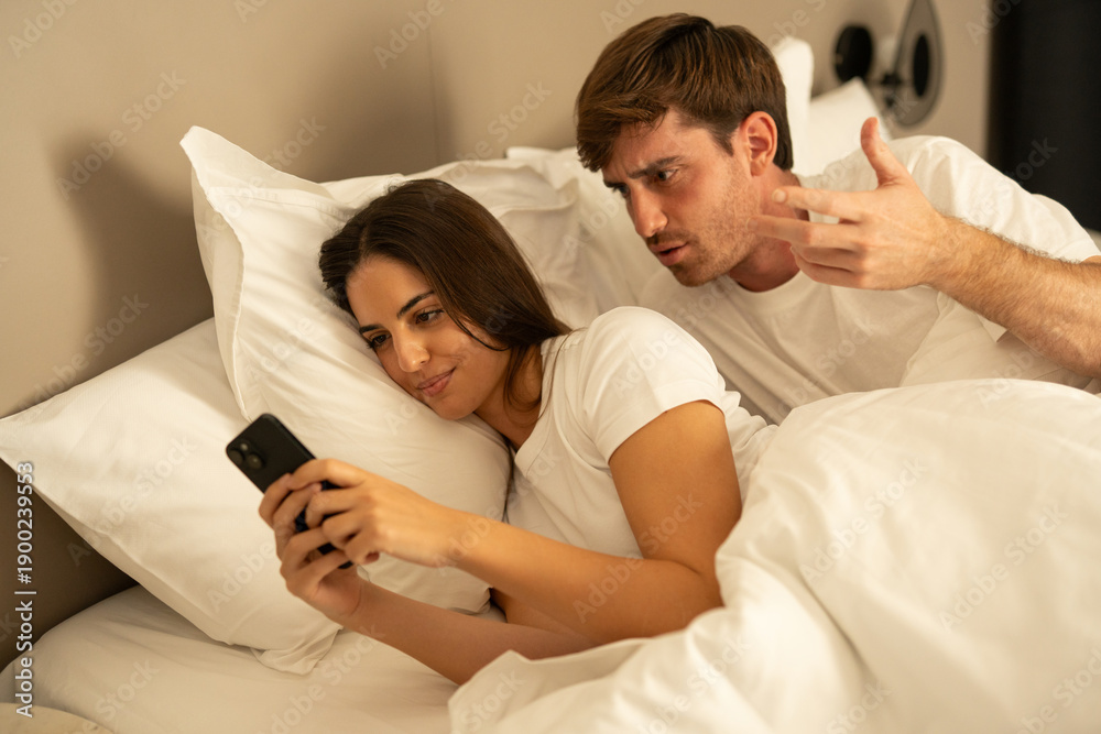 Obraz premium A woman lies in bed, talking on her phone while her partner sleeps soundly beside her. The woman expression suggests she is engaged in a private conversation.