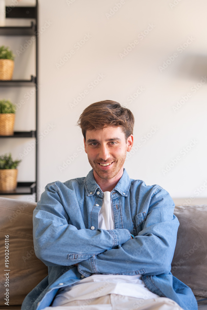 Naklejka premium Young caucasian man indoor sitting on couch smiling and looking at the camera with confidence, crossed arms enjoying the moment. Cheerful guy at home having fun