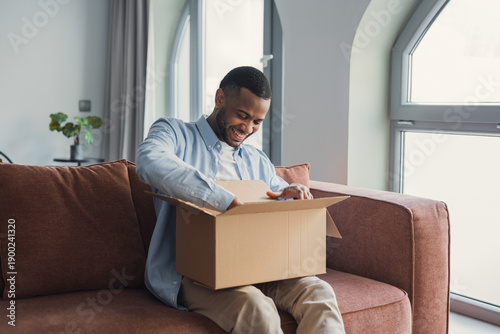 Fotografie Smiling African American cheerful man unpacking parcel, sitting on couch at home