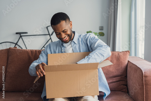 Photos A smiling African American man sits on the couch at home while unpacking a parce
