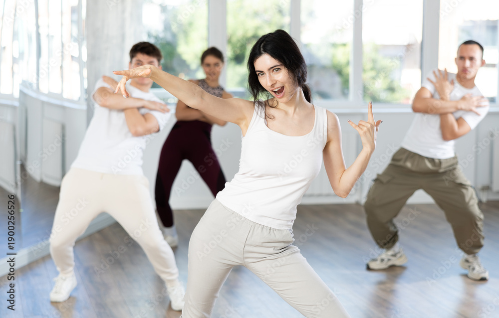 Fototapeta premium Cheerful emotional sporty young female dancer rehearsing popping techniques during group workout in modern street dance studio..