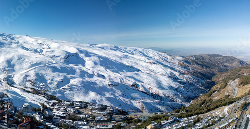 Wide aerial panorama of Sierra Nevada ski resort in winter, showing snowy mountains, village buildings and winding road, ideal background for travel, tourism and winter sports concepts.