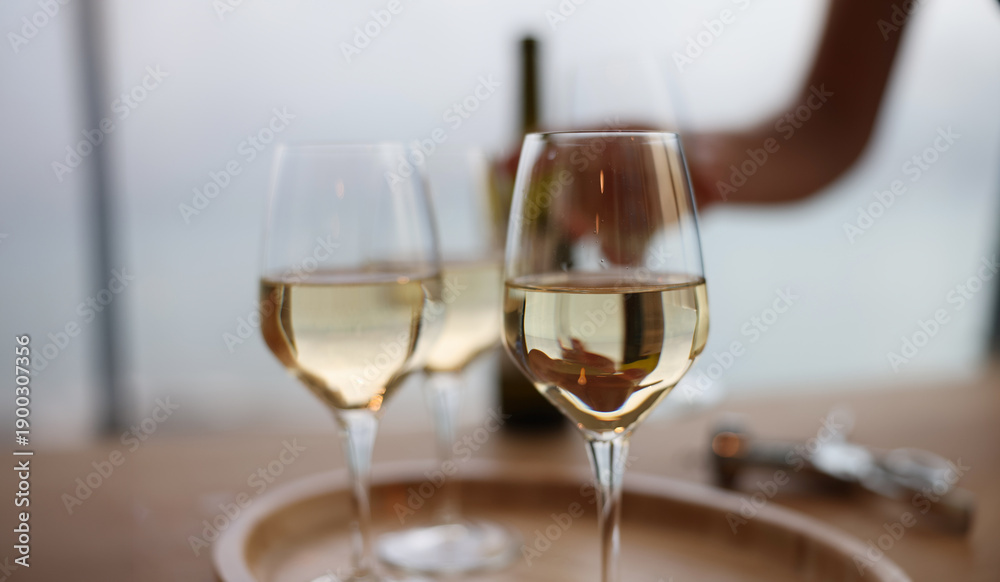 Fototapeta premium Charming white wine glasses sit on a wooden tray, shimmering in the soft light. A gentle hand reaches for the bottle, enhancing the tranquil atmosphere of this midday gathering by the water.