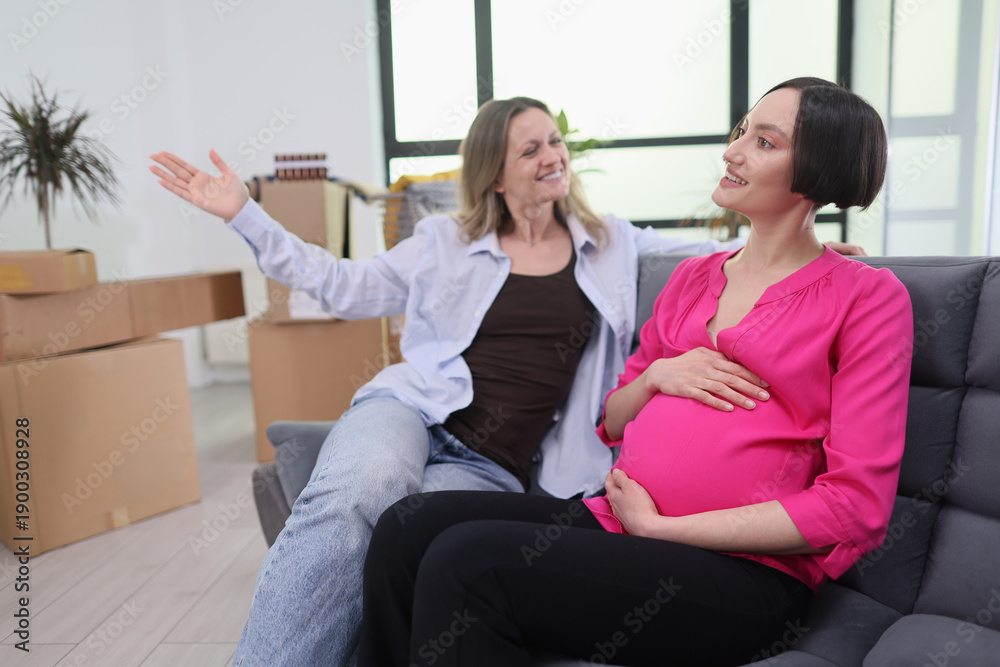 Obraz premium Two friends share a heartwarming moment in a cozy living room. One woman, pregnant and glowing, smiles as the other excitedly gestures, surrounded by moving boxes.