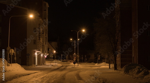 Empty winter street at night illuminated by yellow streetlights. Snow-covered road in a quiet urban residential area.