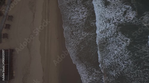 Top-down aerial view of a person walking along the shoreline of Sabaudia beach, Italy. Vertical drone shot of waves crashing on the sand at the foot of Monte Circeo.