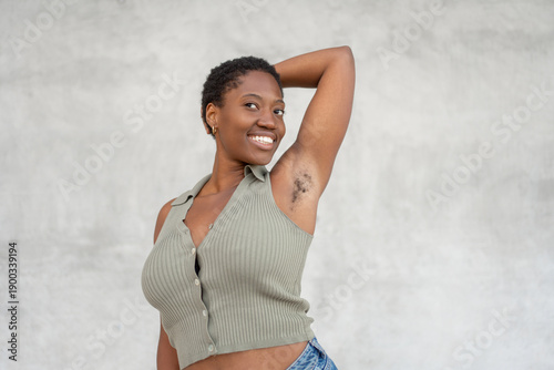 Smiling young woman raising arm and showing underarm hair against gray wall. Body positive and feminism concept, need to shave or depilate