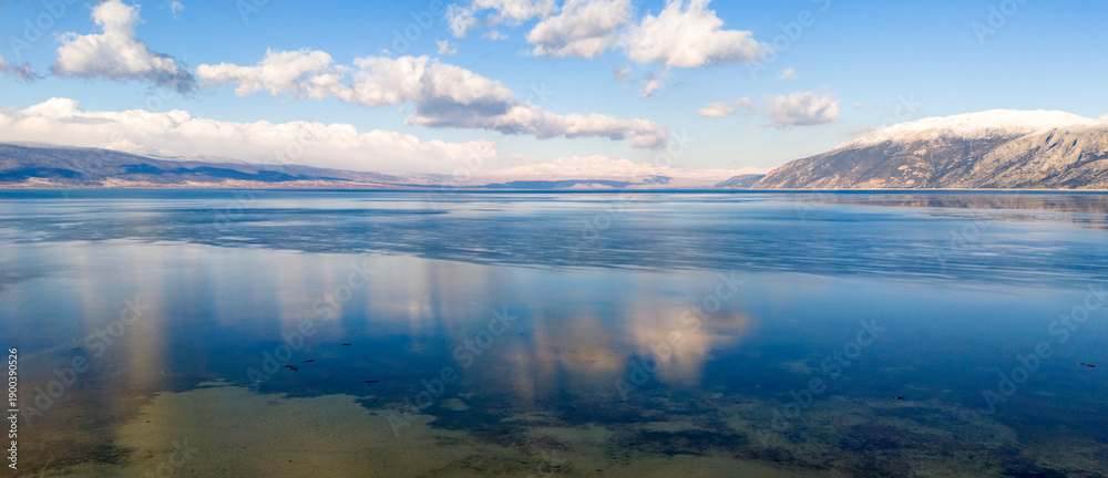 Fototapeta premium Panoramic aerial view of a serene lake with crystal clear water reflecting the blue sky and distant snow covered mountains during winter