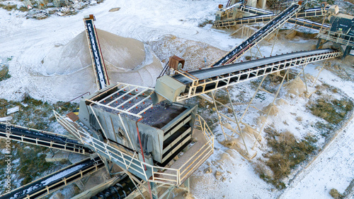 High angle drone shot capturing the intricate conveyor belt infrastructure of an aggregate processing plant during winter. Snow covered ground highlights the heavy mining machinery.