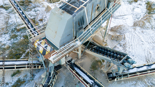 Close-up aerial drone perspective of a massive industrial crushing machine with conveyor belts and caution stripes. Detailed view of heavy mining infrastructure during snowy winter.