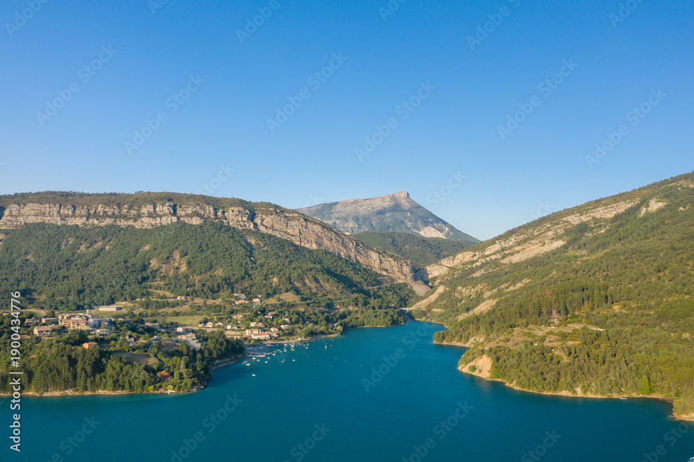 Fototapeta premium Aerial view of a lakeside village nestled among green hills and dramatic cliffs along the turquoise waters of Lac de Castillon, with a prominent mountain peak in the distance and clear summer skies