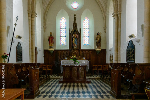 Symmetrical view of a historic church chancel featuring a central altar, flanked by religious statues and tall stained glass windows. Warm wood paneling and checkered tile flooring add texture to the