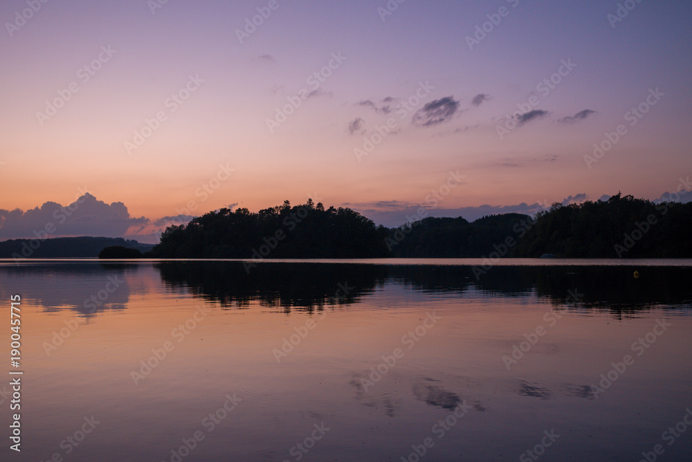 Fototapeta premium Gentle evening light casts soft purple and peach tones across the tranquil surface of Lac des Settons, reflecting the dark outline of distant trees and a serene sky.